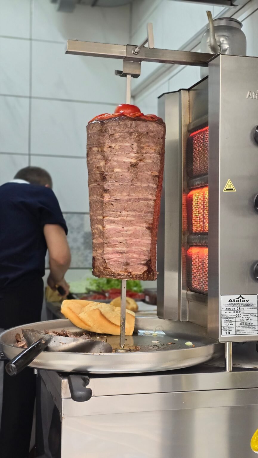 Juicy shawarma meat cooking on a vertical rotisserie with a cook preparing in the background.